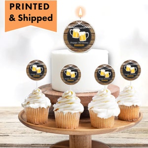 May include: A white cake with a candle and a topper that says "Happy Birthday Brandon!" with a picture of two beer mugs. There are six cupcakes with the same topper on a wooden cake stand.