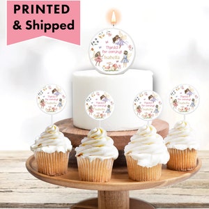 May include: A white cake with a fairy-themed candle topper that says "Thanks for coming Isabella". There are also cupcake toppers with the same design on cupcakes.