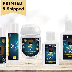 May include: Four different labels for hand sanitizer bottles and lip balm tubes. The labels feature a blue and black space theme with a cartoon boy wearing a virtual reality headset. The text on the labels reads "Noah Reached Level 11" and "Happy Birthday Tristan".