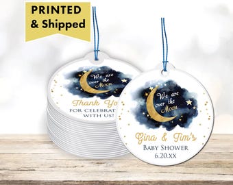 May include: Round white favor tags with blue and gold watercolor designs. The tags feature a crescent moon and stars with the text "We are over the moon" and "Thank you for celebrating with us!"  The other tag has the text "Gina & Tim's Baby Shower 6.20.XX".