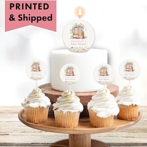 May include: A baby shower cake with a candle and cupcakes. The candle and cupcake toppers feature a floral design with a stack of books and the text "Savannah's Baby Shower". The cupcakes have white frosting.