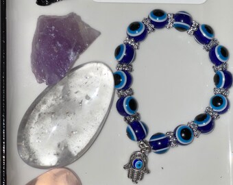May include: A blue and white evil eye bracelet with a silver charm of a hamsa hand with an evil eye. The bracelet is on a white surface with a variety of crystals, including black obsidian, amethyst, clear quartz, and rose quartz.