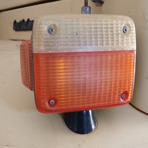 Op de afbeelding: Een close-up van een amberkleurige en witte richtingaanwijzer van een vintage auto. Het licht is rechthoekig van vorm en gemaakt van helder plastic. Er zijn drie zichtbare schroeven die het licht op zijn plaats houden.