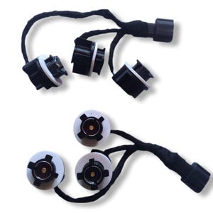 Puede incluir: Vista cenital de varios enchufes de bombillas de automóvil en blanco y negro con cableado. Los enchufes están dispuestos en dos grupos, cada uno conectado a un mazo de cables negro. Los enchufes tienen un diseño circular con un receptáculo central para la bombilla.