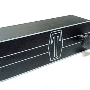 Puede incluir: Caja de metal negro con una perilla plateada y un logotipo plateado de una letra "T" estilizada dentro de un escudo. La caja tiene dos líneas horizontales plateadas.