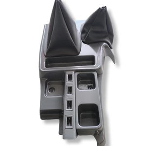 May include: A grey car console featuring two black leather gear stick gaiters. The console includes various compartments, such as cup holders and switch panels. The design is sleek and modern, with a glossy finish.