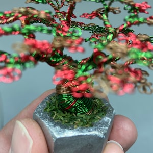 Può includere: Una scultura di albero in miniatura di filo metallico con un tronco e rami in filo metallico verde, rosso e rame. L'albero è in un vaso di metallo argentato con muschio verde.