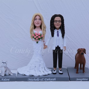 May include: A wedding cake topper set featuring a bride and groom, both with realistic facial features. The bride is wearing a white wedding dress and holding a bouquet of pink flowers. The groom is wearing a white suit and a purple tie. The set also includes two figurines of a cat and a dog.