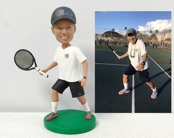 May include: A custom bobblehead figurine of a person playing tennis. The figurine is wearing a white shirt, black shorts, and a grey baseball cap. The person is holding a tennis racket and is in a tennis playing pose. The figurine is standing on a green base.