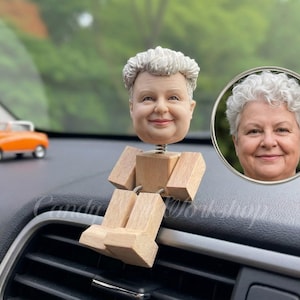 Op de afbeelding: Een op maat gemaakte bobblehead met een lachend gezicht en grijs haar, gemonteerd op een houten lichaam. De bobblehead staat op het dashboard van een auto, met een klein oranje automodel op de achtergrond. Een ingelijst portret van het gezicht is ook zichtbaar.