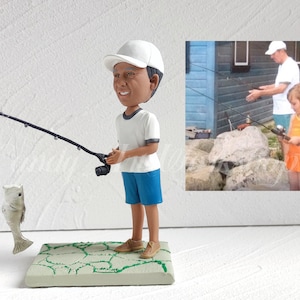 May include: A custom figurine of a person wearing a white hat and blue shorts holding a fishing rod with a fish on the line. The figurine is standing on a green and white base. A photo of a person fishing is in the background.