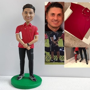 May include: A custom bobblehead figurine of a person wearing a red polo shirt with white trim, black pants, and black shoes. The figurine is holding a white notepad in their left hand and has a watch on their right wrist. The figurine is standing on a green base.