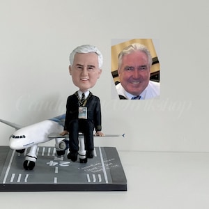 May include: A pilot bobblehead doll sits on a miniature airplane model. The doll is wearing a black uniform with a white shirt and a pilot's tie. The airplane is white and blue, and the base is gray with runway markings.
