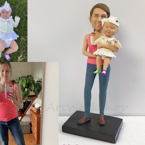 May include: A custom-made figurine of a woman holding a baby. The woman is wearing a pink tank top and blue jeans. The baby is wearing a white dress and a white headband. The figurine is standing on a black base.