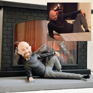 May include: A custom figurine of a person in a black shirt and grey pants, posed in a relaxed position in front of a black fireplace with a faux fire.