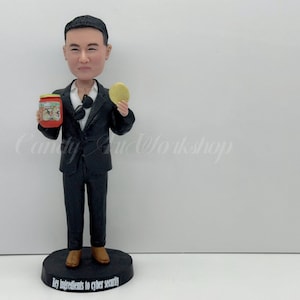 Puede incluir: Figura de un hombre con traje negro sosteniendo un tarro y un disco amarillo. El tarro tiene una etiqueta roja. La figura está sobre una base negra con el texto "Key ingredients to cyber security".