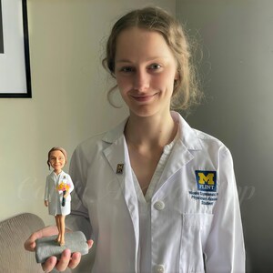Op de afbeelding: Een beeldje van een vrouwelijke dokter in een witte jas, met bloemen, staat op een grijze basis. De persoon die het beeldje vasthoudt, draagt een witte laboratoriumjas met een universiteitslogo en naamplaatje.