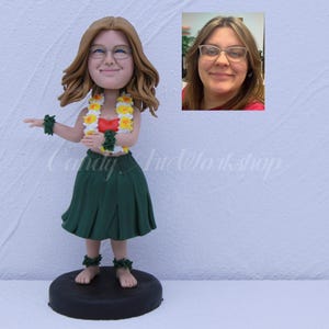 May include: A custom figurine of a person wearing a green hula skirt, a red top, and a white flower lei. The figurine is standing on a black base.