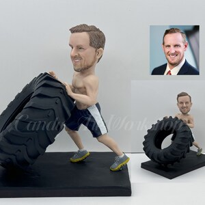 Op de afbeelding: Een op maat gemaakt beeldje van een persoon die een grote band duwt. Het beeldje is gemaakt van geverfd klei en is een realistische weergave van een persoon die aan het sporten is. Het beeldje staat op een zwarte basis.