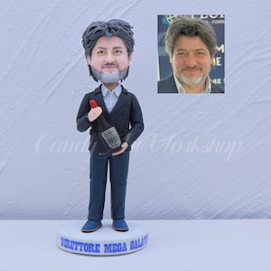 May include: A custom-made figurine of a person holding a bottle of wine. The figurine is wearing a dark blue suit and has a realistic face. The base of the figurine says "Direttore Mega Gala".