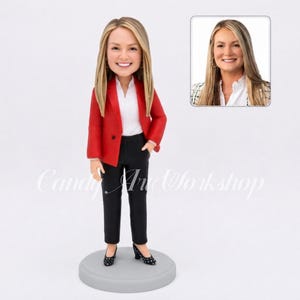 May include: A custom bobblehead figurine featuring a person with long blonde hair, wearing a bright red blazer, a white collared shirt, and black trousers. The figure stands on a circular gray base. A reference photo of the person is included.