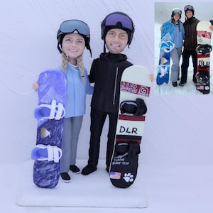 May include: A custom cake topper featuring a couple dressed in winter snowboarding attire, each holding a snowboard. The woman wears a light blue jacket and the man wears a black jacket. The snowboards have the text "DLR" and a paw print.