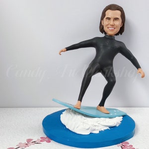 May include: A custom bobblehead figurine of a person wearing a black wetsuit and standing on a blue surfboard with white foam. The figurine is on a blue base with white foam.