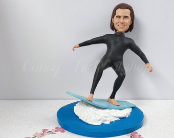 May include: A custom bobblehead figurine of a person wearing a black wetsuit and standing on a blue surfboard with white foam. The figurine is on a blue base with white foam.