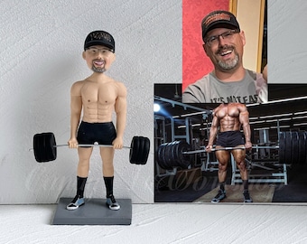 May include: A custom figurine of a person lifting weights in a gym. The figurine is made of painted ceramic and is wearing a black hat, black shorts, and black shoes. The figurine is holding a barbell with weights on each end.