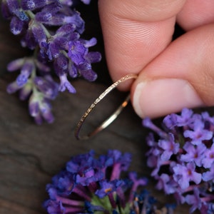 K&ouml;nnte beinhalten: Ein d&uuml;nner Goldring mit einer strukturierten Oberfl&auml;che. Der Ring wird von einer Hand gehalten und ist von lila Blumen umgeben.