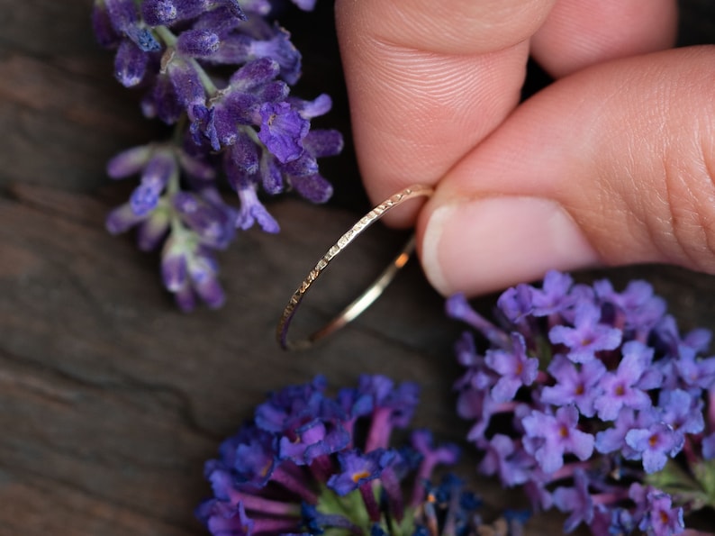 K&ouml;nnte beinhalten: Ein d&uuml;nner Goldring mit einer strukturierten Oberfl&auml;che. Der Ring wird von einer Hand gehalten und ist von lila Blumen umgeben.