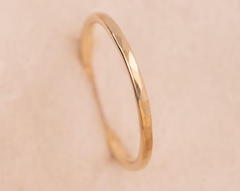 May include: A thin, gold-colored ring with a hammered, faceted texture along the outer surface. The band has a rounded profile and a reflective, metallic finish that catches the light, creating small, distinct highlights across its surface.