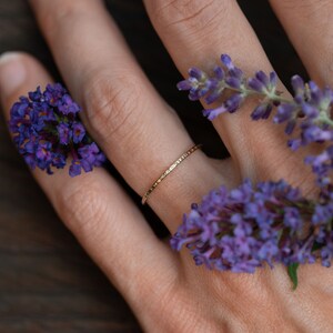 K&ouml;nnte beinhalten: Ein zarter Goldring mit geh&auml;mmerter Textur, getragen an einem Finger, der mit lila Blumen geschm&uuml;ckt ist.