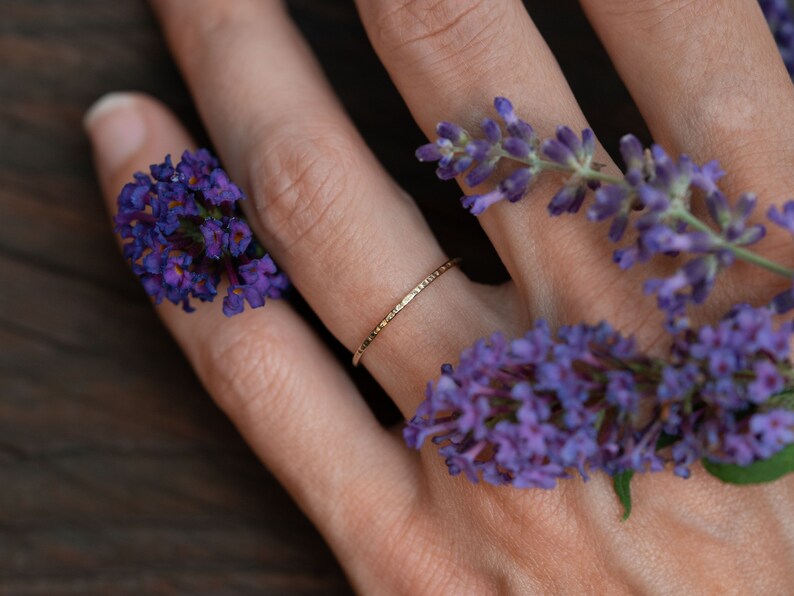 K&ouml;nnte beinhalten: Ein zarter Goldring mit geh&auml;mmerter Textur, getragen an einem Finger, der mit lila Blumen geschm&uuml;ckt ist.