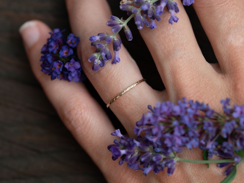 K&ouml;nnte beinhalten: Ein zarter goldener Ring mit geh&auml;mmerter Textur, getragen an einem Finger, der mit violetten Lavendelbl&uuml;ten geschm&uuml;ckt ist.