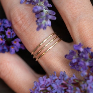 K&ouml;nnte beinhalten: Drei goldene Stapelringe an einem Finger, mit lila Blumen im Hintergrund.