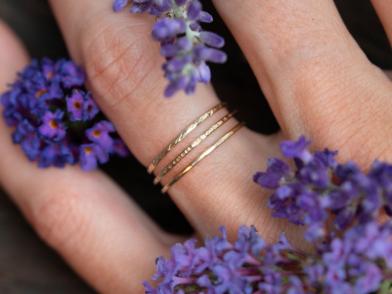 K&ouml;nnte beinhalten: Drei goldene Stapelringe an einem Finger, mit lila Blumen im Hintergrund.