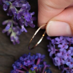 K&ouml;nnte beinhalten: Ein d&uuml;nner Goldring mit geh&auml;mmerter Textur. Der Ring wird von einer Hand gehalten und ist von lila Blumen umgeben.