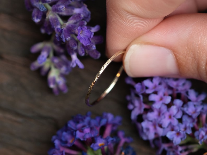 K&ouml;nnte beinhalten: Ein d&uuml;nner Goldring mit geh&auml;mmerter Textur. Der Ring wird von einer Hand gehalten und ist von lila Blumen umgeben.