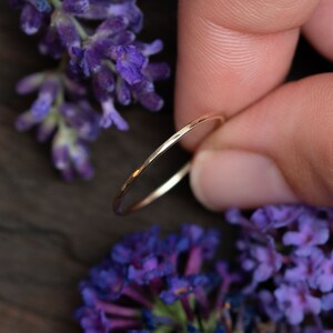 K&ouml;nnte beinhalten: Ein d&uuml;nner Goldring mit geh&auml;mmerter Textur. Der Ring wird an einem Finger mit lila Blumen im Hintergrund gezeigt.