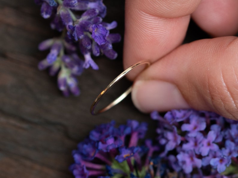 K&ouml;nnte beinhalten: Ein d&uuml;nner Goldring mit geh&auml;mmerter Textur. Der Ring wird an einem Finger mit lila Blumen im Hintergrund gezeigt.