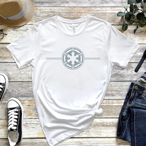 Puede incluir: Camiseta blanca con un símbolo imperial gris de Star Wars en la parte delantera. El símbolo está centrado en la camiseta y tiene una fina línea gris por encima y por debajo.