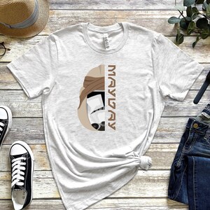 Könnte beinhalten: Ein weißes T-Shirt mit einer braunen und weißen Illustration eines Stormtrooper-Helms mit dem Text "MAY THE 4TH" vertikal auf der rechten Seite des Bildes.