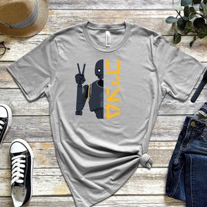 Puede incluir: Camiseta gris claro con un gráfico de robot y el texto "K:2SO" en amarillo. La imagen incluye zapatillas negras, vaqueros y accesorios. La camiseta es una prenda informal.