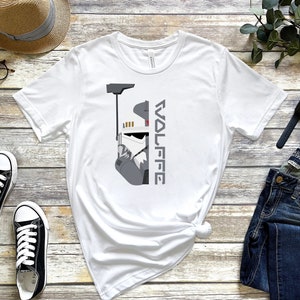 May include: White t-shirt with a grey and white illustration of a Star Wars clone trooper helmet and the text "WOLFFE" printed on the right side of the shirt.