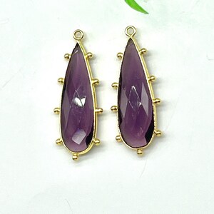 Puede incluir: Dos colgantes de amatista en forma de lágrima con marcos dorados y pequeños detalles dorados. Los colgantes se muestran sobre una superficie blanca con hojas verdes en el fondo. Cada colgante tiene un pequeño lazo en la parte superior.