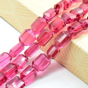 Puede incluir: Tres hilos de cuentas rosas rectangulares facetadas. Las cuentas están ensartadas, mostrando su calidad translúcida. Las cuentas descansan sobre una caja beige con un sutil patrón geométrico.