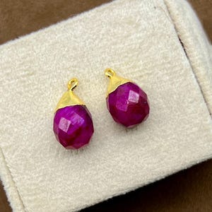 Könnte beinhalten: Zwei facettierte, tropfenförmige Ohrringe in tiefem Magenta. Jeder Ohrring hat eine goldfarbene Kappe. Die Ohrringe werden auf einer cremefarbenen, rechteckigen Schmuckschatulle vor einem braunen Hintergrund präsentiert. Schmuck.