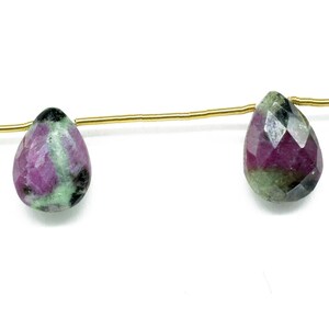 Może przedstawiać: Dwie koraliki w kształcie łzy, wykonane z kamieni szlachetnych, nawleczone na złoty drut. Koraliki są połączeniem zieleni, różu i czerni.