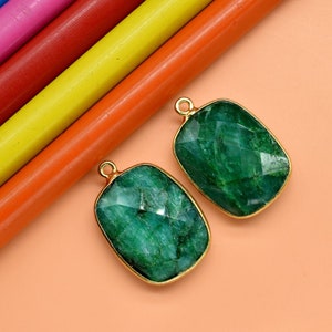 May include: Two green gemstone pendants with gold settings. The gemstones are rectangular and have a faceted cut. The pendants are on a light orange background.